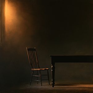 A single wooden chair drawn up to a bare table in a dim room, the seat empty.
